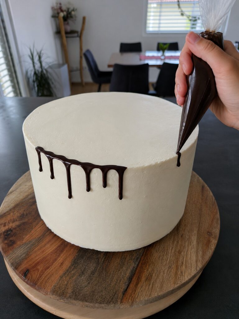 Schokoladen-Drip wird mit einem Spritzbeutel auf eine weiß eingestrichene Torte aufgetragen.